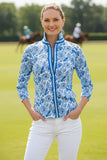 Woman wearing a blue and white patterned blouse on a grassy field with polo players and horses in the background.